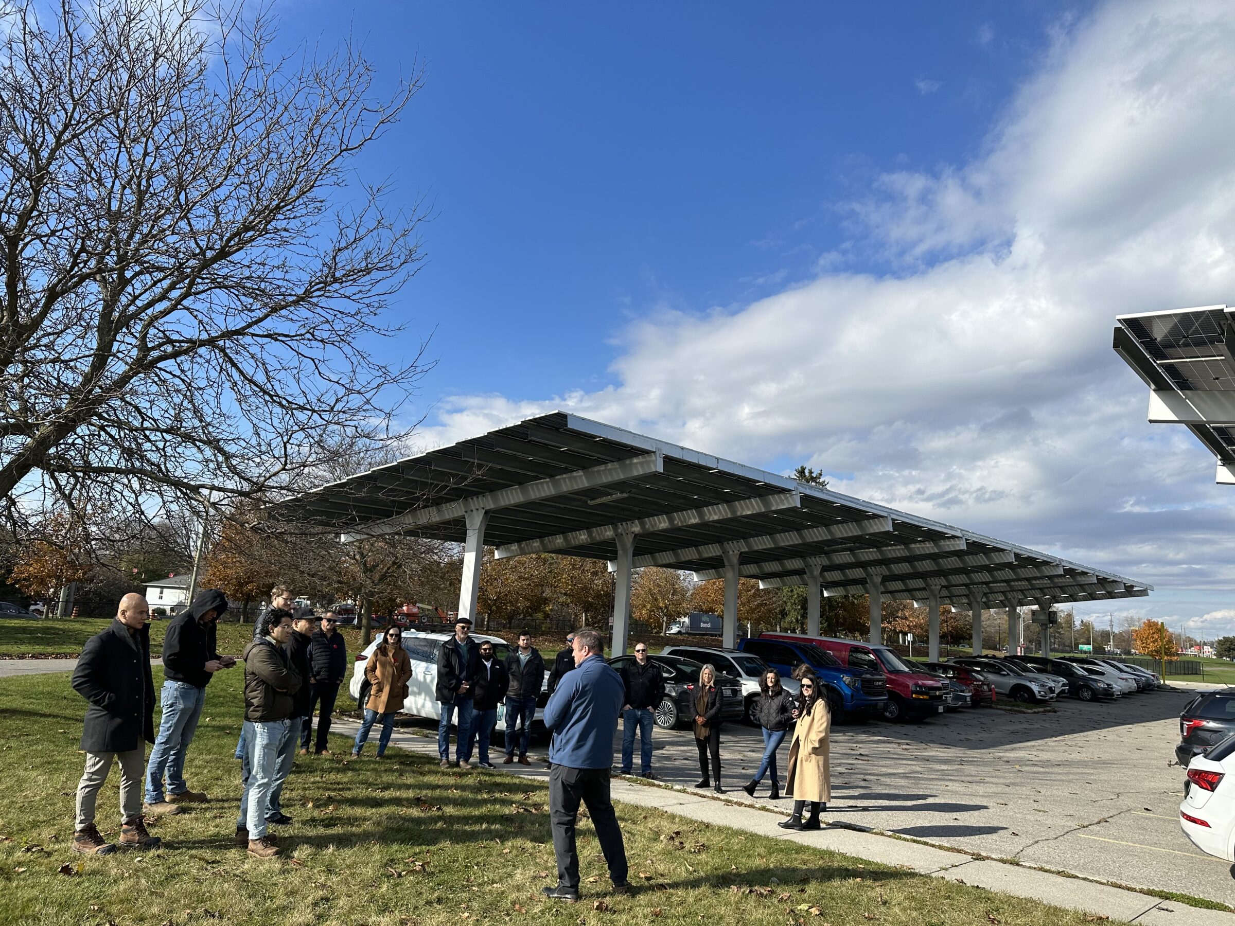 A group of people gather near parked cars under solar panel canopies, with a clear blue sky and bare trees in the background.