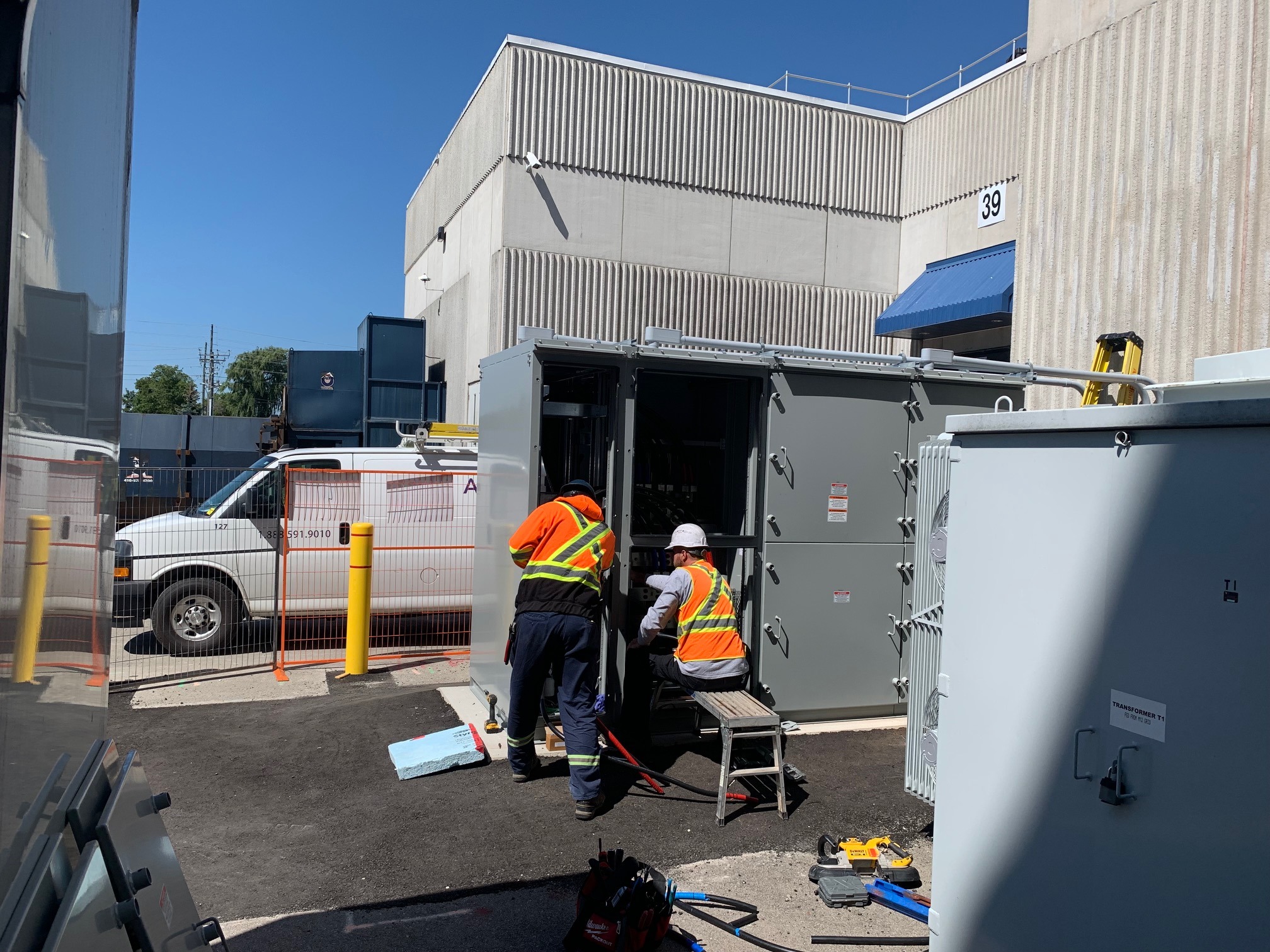 Two people in safety gear work on electrical equipment outside an industrial building. A white van is parked nearby under a clear sky.