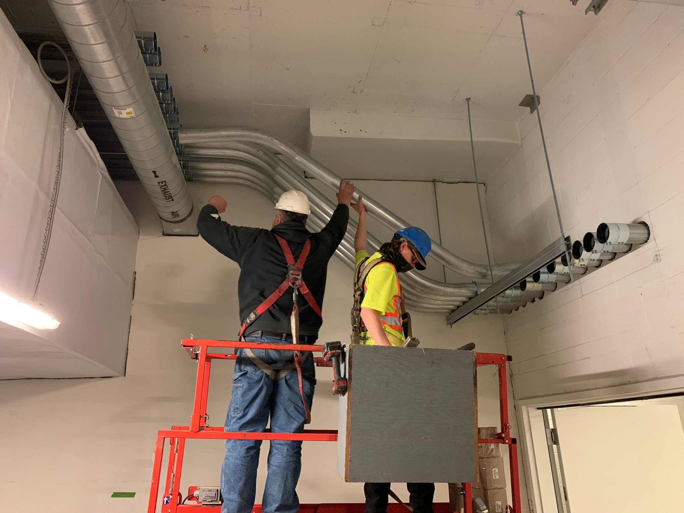 Two people on a lift work on installing large pipes in an industrial indoor setting. Safety gear and tools are visible around them.