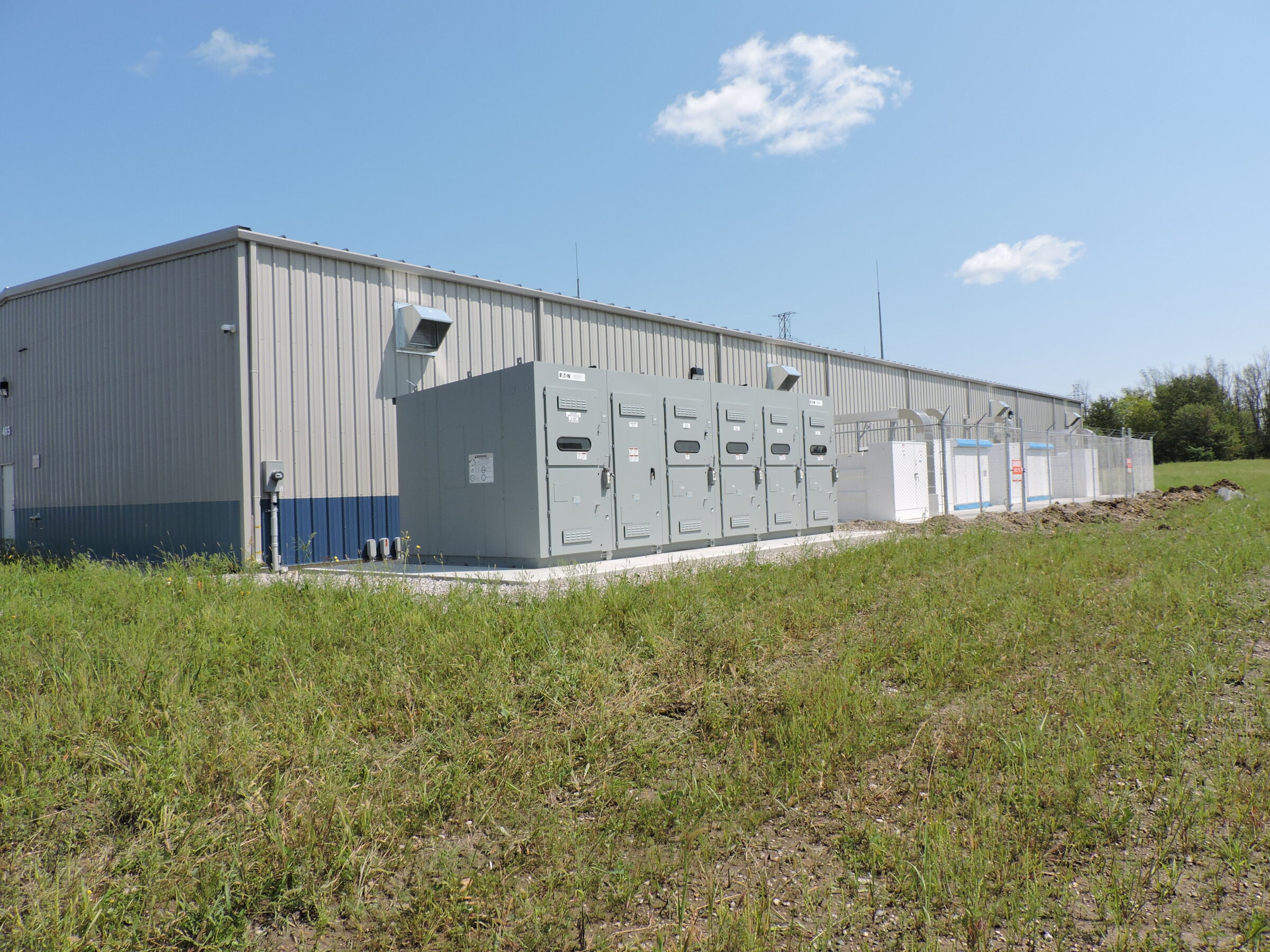 A large industrial building with metal cladding sits beside an outdoor utility area, surrounded by grass, under a clear blue sky with clouds.
